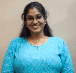 Woman in a light blue patterned top wearing glasses, smiling at the camera against a plain light-colored background.