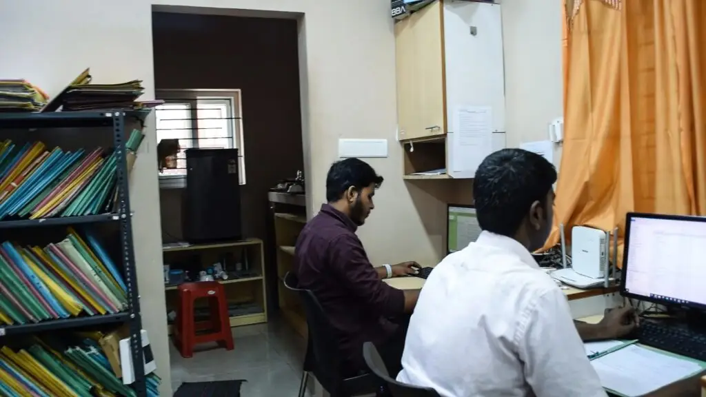 Two members of the K. Ramanan & Co team working at computers in a compact office, with shelves of colourful files in the background.