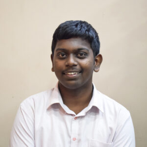 Young man in a light pink shirt smiling at the camera against a plain light-colored background.