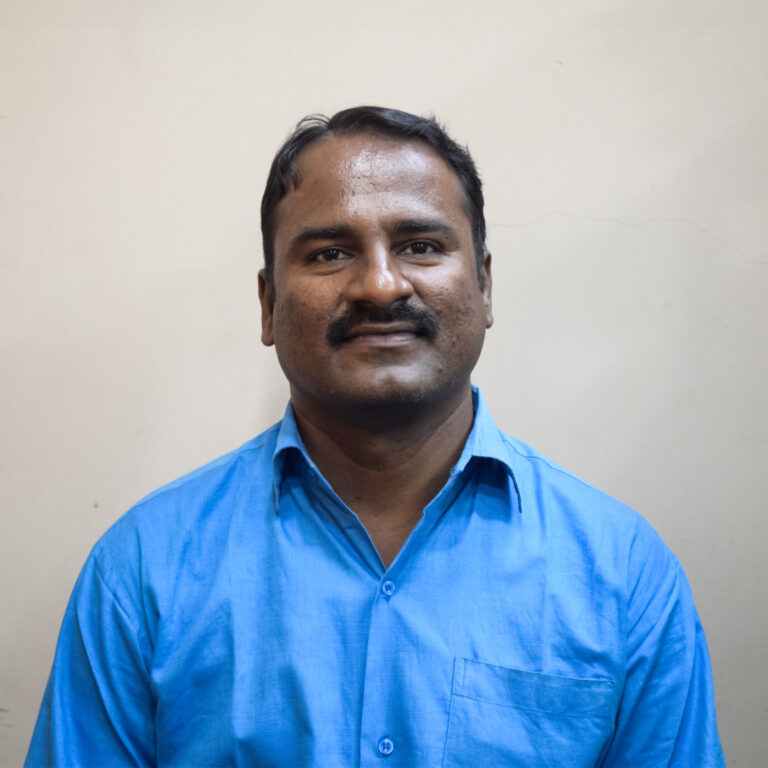Man in a bright blue shirt standing against a plain light-colored background, looking directly at the camera with a slight smile.