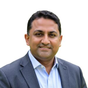 Professional headshot of Ajoy Ramanan wearing a dark blazer and light blue shirt, standing against a plain white background